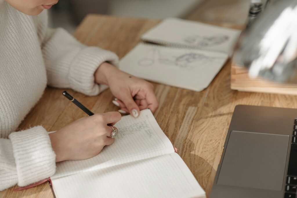 A woman writes in a lined notebook at a wooden desk with a laptop and sketchbook nearby.